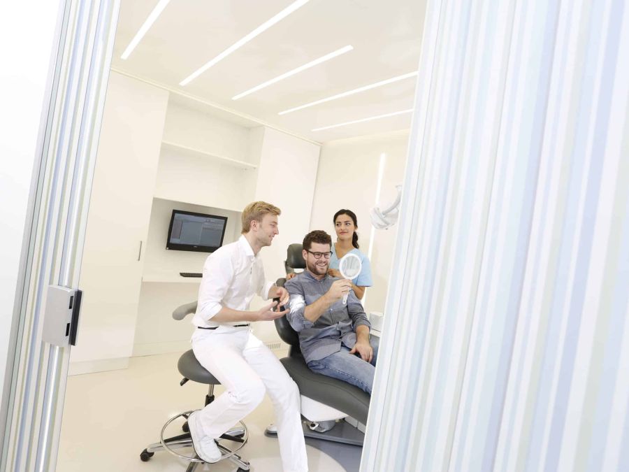 Zahnarzt Dr. Thomsen, ein Patient und eine Mitarbeiterin im Behandlungsraum. Der Patient hält einen Spiegel in der Hand und betrachtet das Behandlungsergebnis.